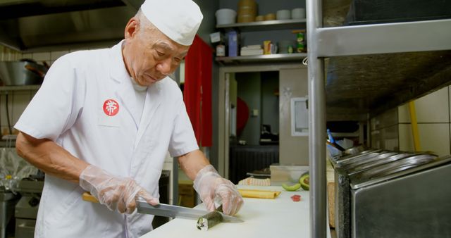 This image captures an experienced sushi chef skillfully slicing fresh fish on a kitchen counter in a restaurant setting. The chef is wearing a traditional uniform, including a white hat and gloves, while meticulously preparing sushi. The kitchen environment highlights his dedication and expertise in Japanese cuisine. This visual can be used for content related to culinary arts, Japanese food, professional cooking, and restaurant promotions.