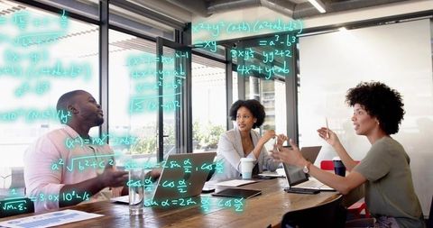 Team collaborating on teal math equations on glass wall around wooden table with laptops
