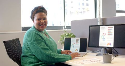 Smiling african american woman typing on laptop with colorful charts on dual screens