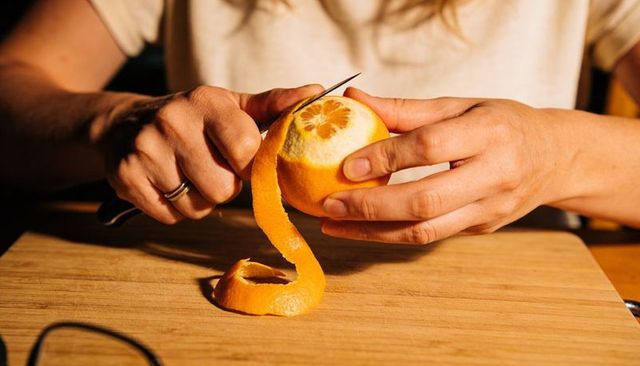 Peeling orange with paring knife, hands curling long citrus peel on wooden cutting board