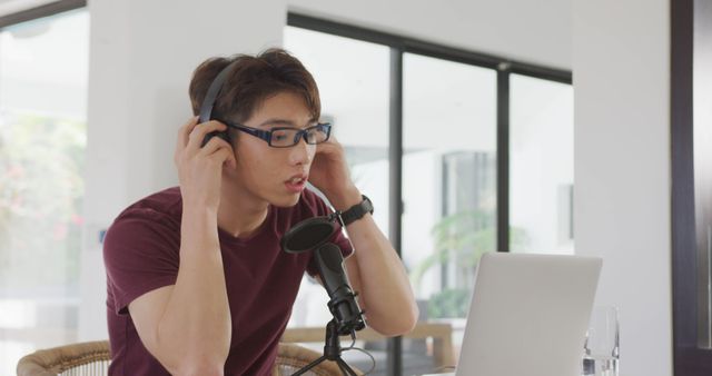 Young Asian man recording a podcast at home. He is adjusting his headphones, speaking into a microphone, and using a laptop. Ideal for use in articles or posts about podcasting, remote work, technology, home studios, digital content creation, and modern communications.