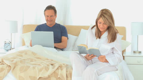 Mature couple enjoys quiet morning routine in bright bedroom. Man using laptop while woman reading book. Peaceful start to the day ideal for illustrating balanced lifestyle and morning routines in promotional material.