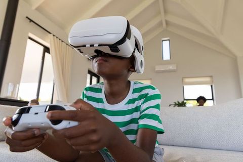 African american boy wearing vr headset playing with controller on bright modern couch