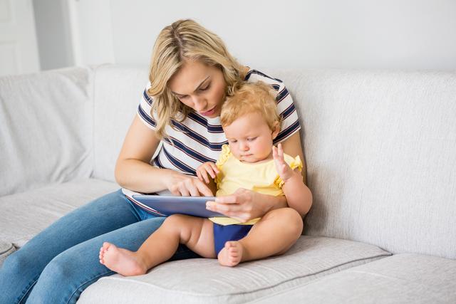 Mother and baby girl sitting on sofa using digital tablet together. Ideal for content related to family bonding, parenting tips, early childhood education, and technology use in family settings.