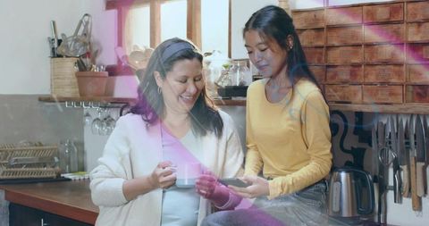 Cozy morning sharing smartphone and coffee: mother and daughter smiling in sunlit kitchen