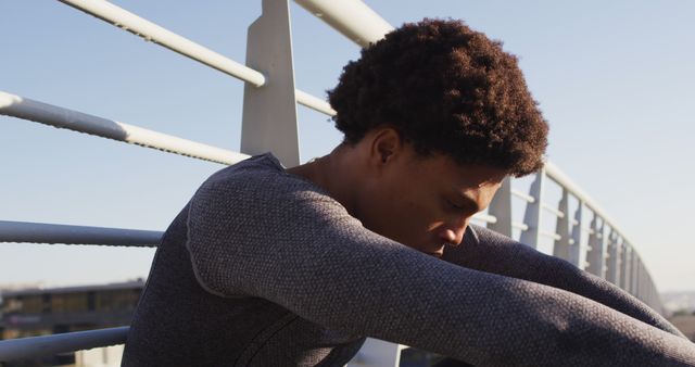 A young man with black hair leans on a bridge, lost in deep thought under the bright sun. This visual conveys themes of solitude, introspection, and personal reflection. Ideal for articles about mental health, lifestyle blogs focusing on mindfulness and meditation, or marketing materials promoting outdoor activities and personal growth.