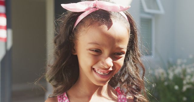 A cheerful young girl wearing a pink bow in her hair is enjoying a sunny day outdoors. Her smile radiates joy under the natural sunlight. Can be used in contexts related to childhood, happiness, summer activities, and fashion for children.