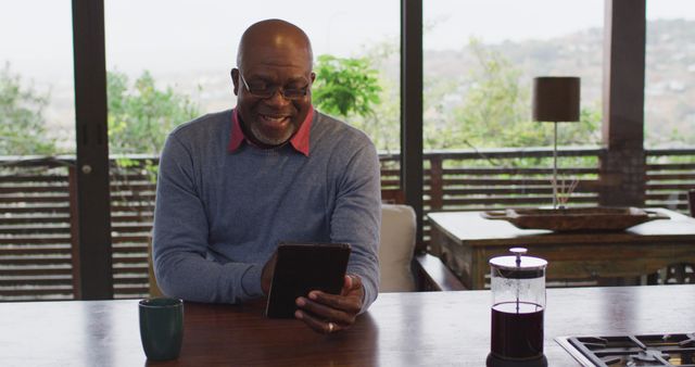 A senior man is smiling while using a digital tablet at a wooden table during the morning at home. There is a cup and a French press on the table, and the background features large windows revealing an outdoor area with greenery. This image can be used for articles on technology use among older adults, lifestyle blogs, retirement living, and advertisements for home technology products.