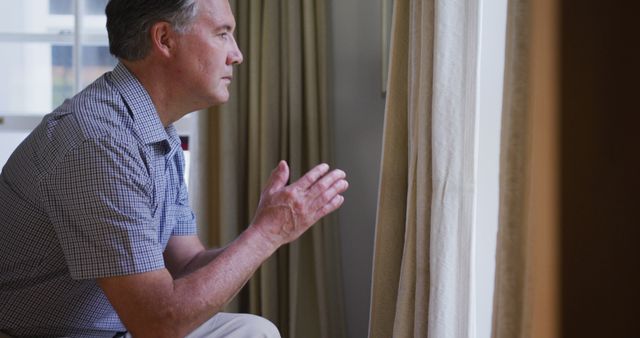 Mature man sitting indoors, contemplating while looking through window. Best for themes like reflection, thoughtful moments, introspection and solitude. Useful for mental health articles, blogs about aging and personal growth.