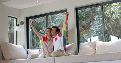 Joyful Gay Couple Holding Rainbow Flag Sitting with Smartphone