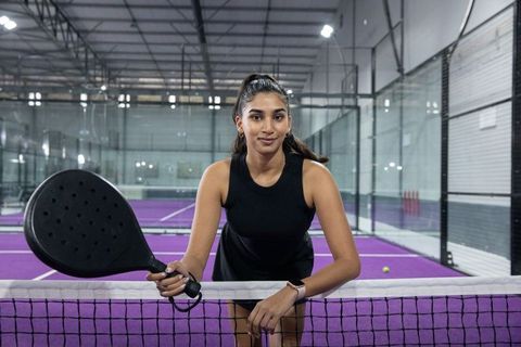 Indian woman playing padel on purple indoor court leaning on net holding racket