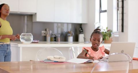 African american mom supervising daughter doing homework at kitchen table with laptop