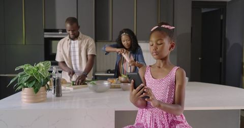African American family cooking together in modern kitchen while daughter using smartphone
