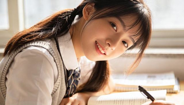 Smiling student in school uniform leaning at sunlit desk writing in open textbook with pen