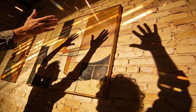 Shadow figures reaching toward abstract geometric painting on sunlit brick wall