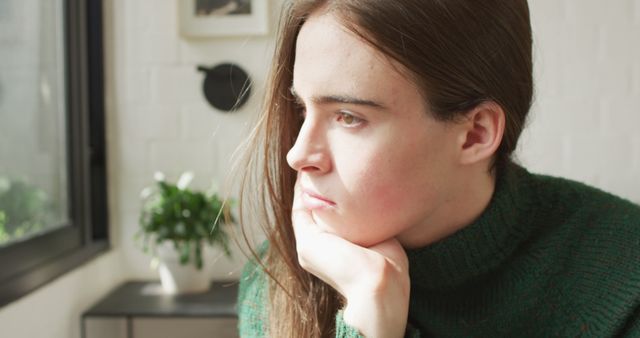 Young woman wearing a green sweater is looking thoughtfully out the window in a cozy indoor setting. Natural light is illuminating her face, giving a calm and serene atmosphere. Useful for themes related to thinking, contemplation, mindfulness, calmness, solitude, or indoor lifestyle.