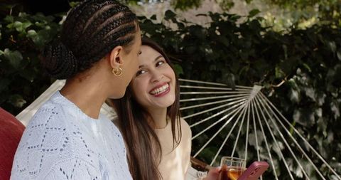 Two friends laughing on hammock while sharing smartphone and iced drink in backyard garden