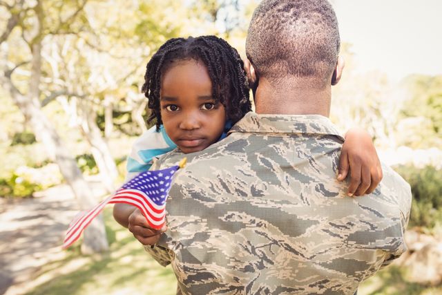 Military father holding his young daughter who is holding an American flag. They are outdoors in a park, showcasing family bonding and patriotism. Ideal for use in campaigns promoting military family support, patriotic events, and family values.