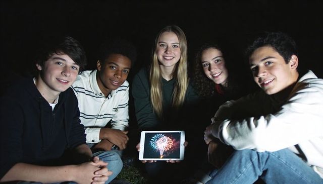 Teen friends gathering at night holding tablet showing fireworks celebrating together