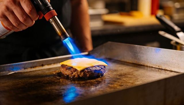 Chef torching burger patty with blowtorch melting cheese on hot griddle
