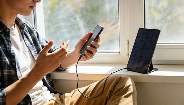 Man charging smartphone with portable solar panel on windowsill, sustainable lifestyle