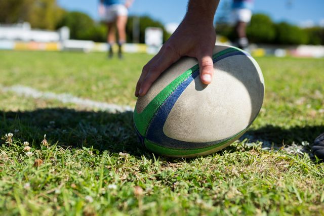 Close-up of a player's hand touching a rugby ball on a grassy field. Ideal for use in sports-related content, team spirit promotions, athletic training materials, and outdoor activity advertisements.