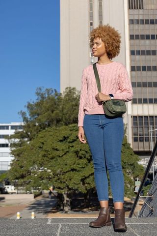 Sunlit urban woman standing on plaza ledge wearing pink sweater, denim, green crossbody