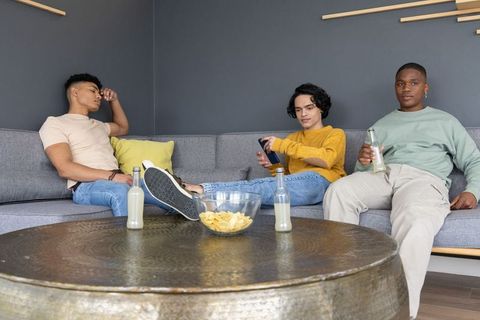 Multiethnic young men relaxing on modern sofa sharing drinks and chips in minimalist lounge