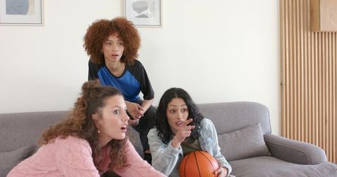 Multicultural women sitting on couch holding basketball and pointing while watching game