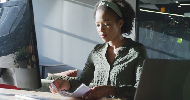 Young woman sitting at desk in modern office setting, focused on holding and organizing documents. Ideal for illustrating themes of professionalism, business environments, modern workplaces, productivity, and technology use in offices.