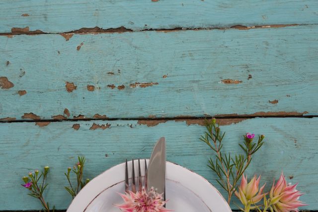 Overhead view of a rustic table setting featuring cutlery and flowers on a white plate. Ideal for use in dining, home decor, and lifestyle blogs. Perfect for illustrating concepts related to rustic or vintage dining experiences, floral arrangements, and table decor.