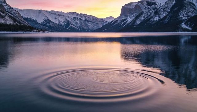 Alpenglow sunrise over snowy mountains reflecting on calm lake with rippling rings at dawn