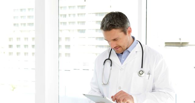 Male doctor in white coat and stethoscope using a digital tablet in a bright room. He is concentrating on the device, suggesting the use of modern technology in healthcare services. Ideal for use in articles or advertisements regarding modern healthcare, technology in medicine, medical consultations, professional medical services, and healthcare settings.