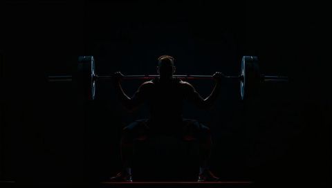 Squatting male lifter holding olympic barbell in dramatic low-key gym lighting