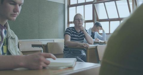 Contemplative student wearing glasses and striped tee resting chin at desk with notebooks