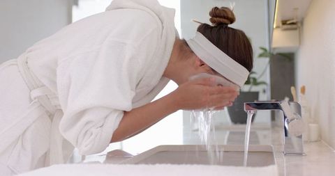 Woman splashes water on face wearing bathrobe in contemporary bathroom