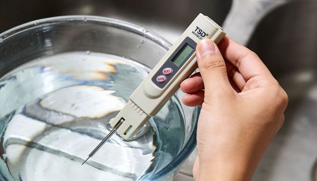 Right-hand holding handheld tds meter testing water quality in glass bowl at kitchen sink