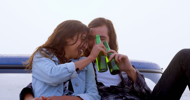 Young couple cheerfully drinking beers while sitting at beach after sunset. Ideal for depicting relaxation, vacation moments, leisure activities, or romantic getaways. Suitable for ads promoting beach resorts, beverages, or travel experiences.