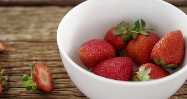 Colorful ripe strawberries are placed in a sleek white bowl on a rustic wooden table. This setting creates a contrast, highlighting the red berries. Ideal for use in promoting healthy eating, organic produce, summer recipes, or stylish foodie blogs.