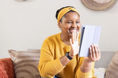 African american woman waving during tablet video call on cozy sofa wearing mustard sweater