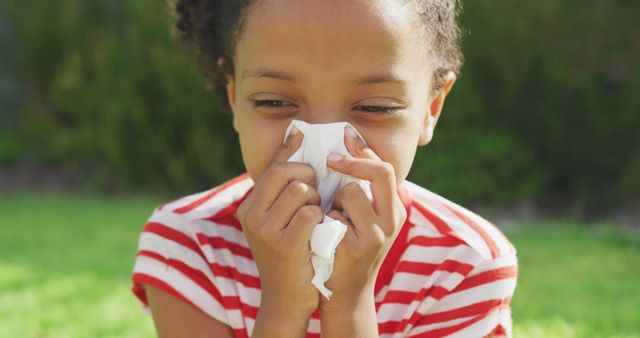 Young girl with curly hair using a tissue to blow her nose while sitting outside on a sunny day. Employ this image for articles about seasonal allergies, cold and flu awareness, children's health, or outdoor play during allergy season.