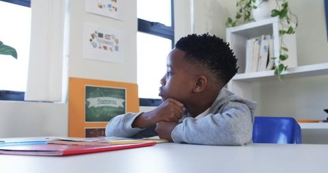 Pensive african schoolboy sitting at desk, looking out window in modern classroom light