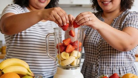 Friends preparing strawberry banana smoothie in blender on bright home kitchen counter