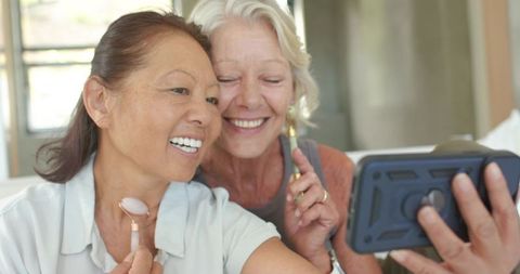 Mature asian and caucasian women sharing skincare ritual and taking joyful selfie on couch