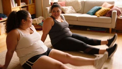 Curvy women stretching together in cozy living room while checking animated fitness stats