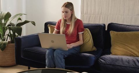 Young woman working on laptop on navy velvet sofa in cozy modern living room