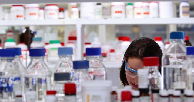 Image of a female scientist wearing safety goggles focusing on her research amidst various chemicals and laboratory bottles in the background. Suitable for articles or advertisements related to scientific research, laboratory work, female scientists in STEM, or educational purposes highlighting laboratory procedures and scientific experiments.