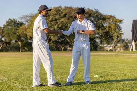 Cricket teammates shaking hands on sunlit field wearing whites and black caps during training
