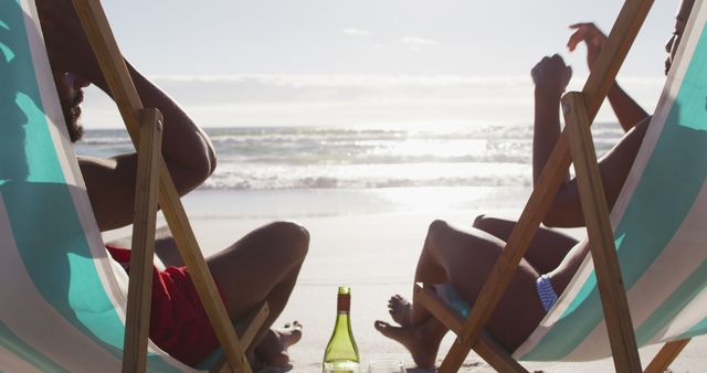 Friends sitting in deck chairs enjoying a relaxed evening at the beach. A bottle of beverage is placed in the sand nearby. Ideal for concepts related to relaxation, vacation, summer beach outings, friendship, and leisure activities.