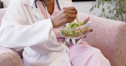 Female nurse sitting on couch in staff lounge eating salad during break with stethoscope